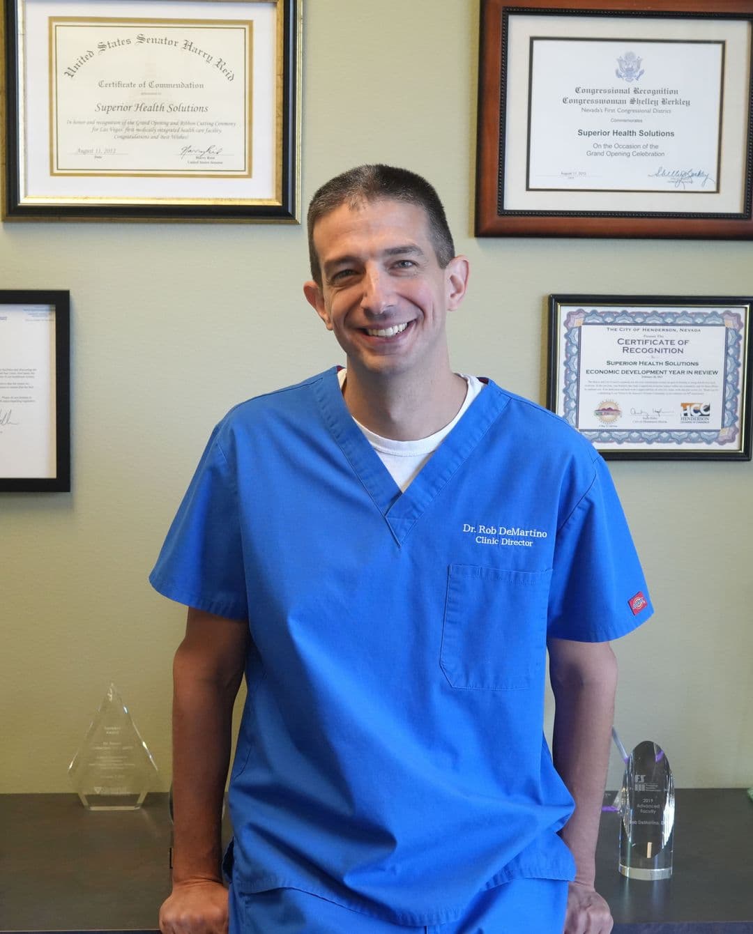 Dr. Rob DeMartino in clinical scrubs at Superior Health Solutions, standing in front of framed community recognition awards